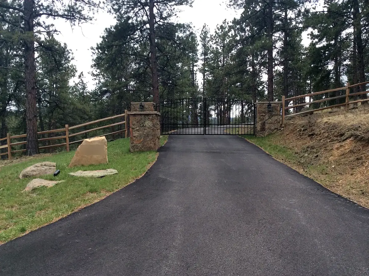 Asphalt driveway rising to an iron gate between stone pillars, with split-rail fence and boulders, Evergreen, Colorado