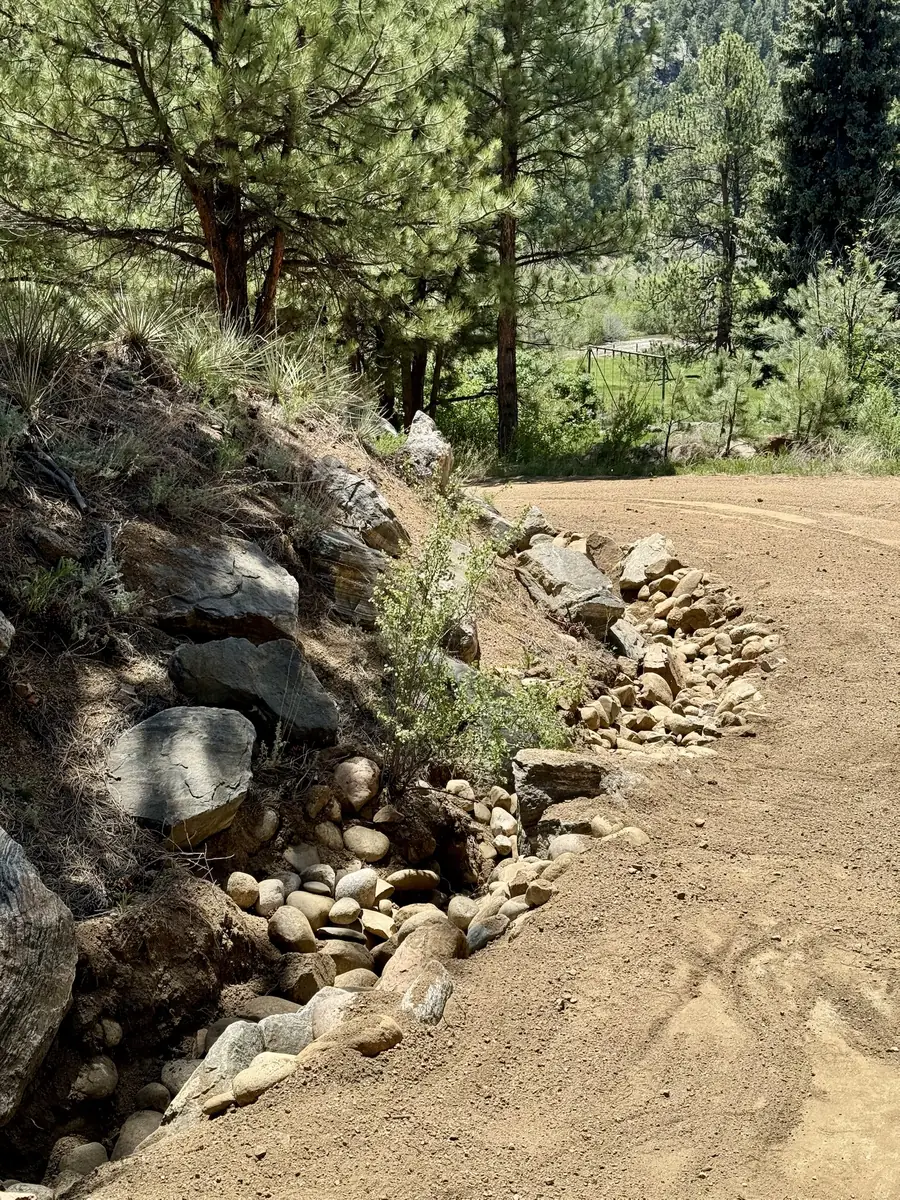Rock-lined drainage ditch curving along the uphill edge of a regraded gravel mountain driveway under ponderosa pines