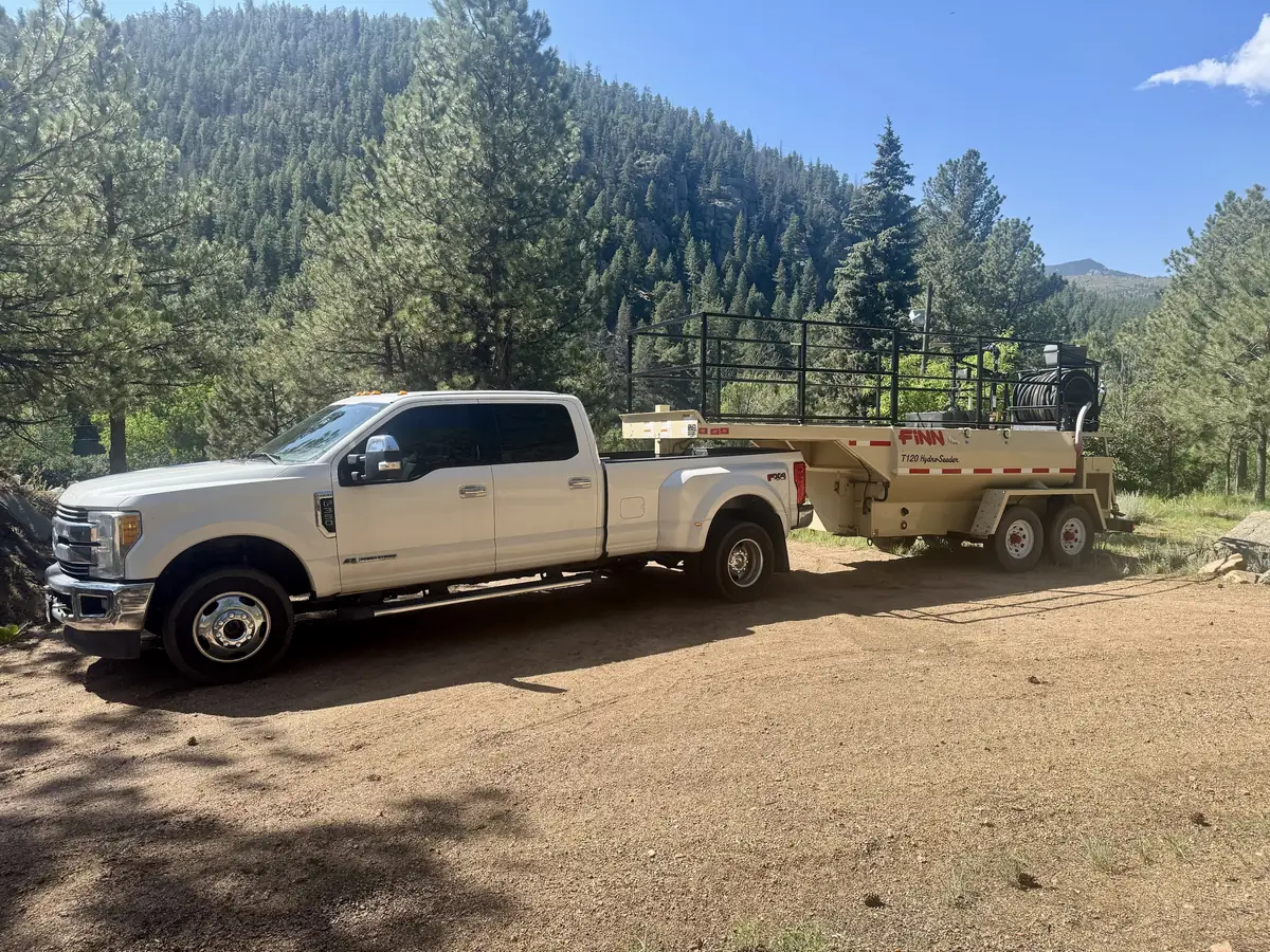 White Ford F-350 dually truck towing a hydroseeder trailer on a dirt mountain road with pine-covered hills behind